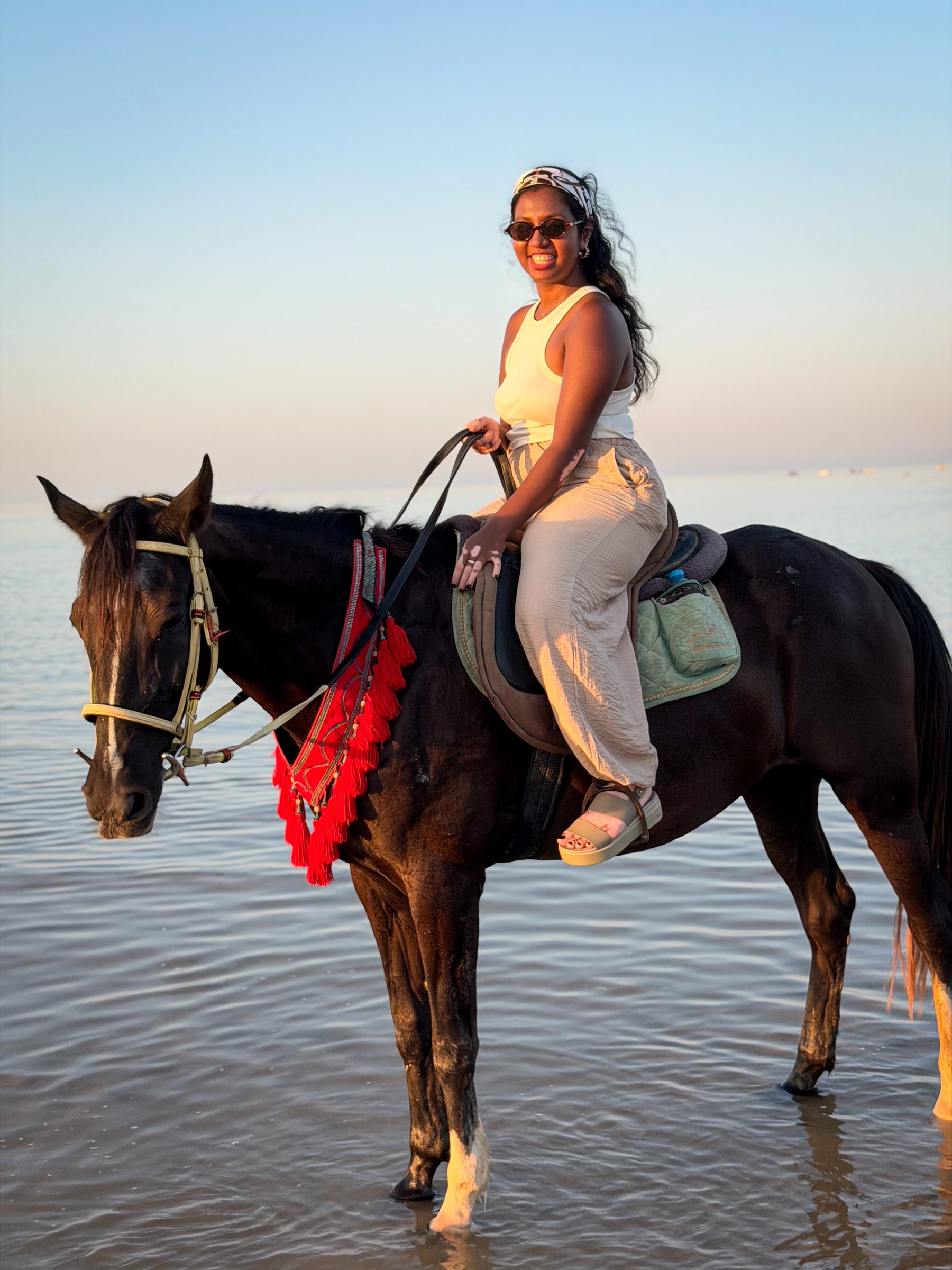 Priya smiling on horseback in shallow sea water at sunset.