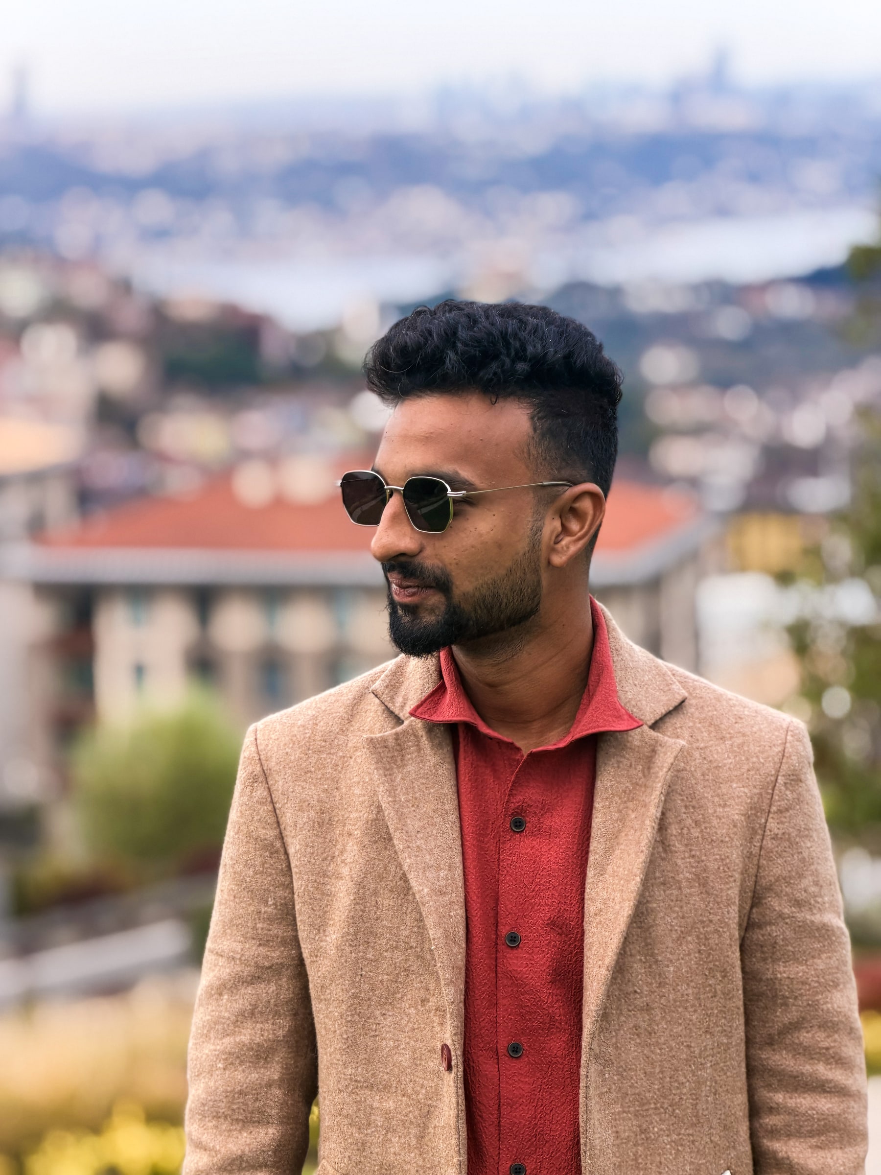 Santhosh in sunglasses and a camel blazer, looking out over a city.